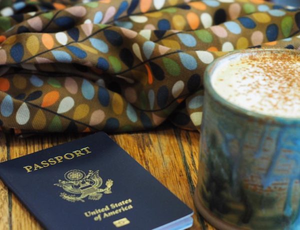 A picture of my first United States Passport with an almond milk latte in a ceramic mug beside it and a scarf with softly colored stylistic leaves on it in the background