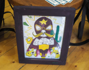 Picture shows an illustration called The Wrestler by Little Rocks Design. It is a wrestler with tattooed arms in a yellow and purple leotard, a purple cape and tights, a big yellow handlebar mustache, and a purple full face mask with yellow star on the forehead. Surrounding him are three bull terriers wearing miniature yellow masks and costumes to match the wrestler.