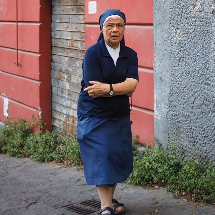 An old Catholic Nun in her blue dress, sweater, habit and white shirt, walking down a quiet alley against a backdrop of red and grey colored stone buildings in Naples Italy.